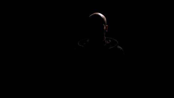Ο φαλακρός με τη μαύρη κουκούλα κοιτάει στην κάμερα πριν σηκώσει την κουκούλα του. Το πρόσωπό του σκιάζεται από το πίσω φως, το οποίο δημιουργεί μυστηριώδη διάθεση. Απομονωμένο πορτραίτο ενός ξένου. Αίσθηση μυστηρίου και ίντριγκας. - Πλάνα, βίντεο