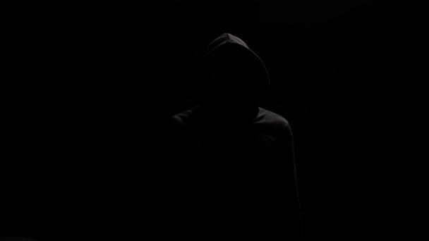 Figura misteriosa retrato aislado en sudadera con capucha negra, apuntando al dedo de la cámara. La figura del hombre con capucha negra emerge de las sombras, dirigiendo el dedo apuntando a la lente. Desconocida vestida de negro en retroiluminación - Imágenes, Vídeo