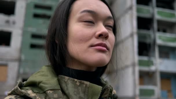 Close-up retrato de menina asiática em uniforme forças armadas ucranianas vira a cabeça e olha para edifício residencial destruído durante o bombardeio pelo exército russo. Batalhão militar internacional na Ucrânia - Filmagem, Vídeo