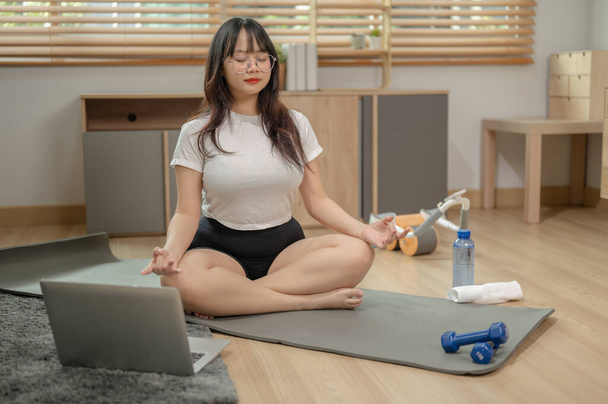A calm, relaxed young Asian woman in sportswear is meditating on a yoga mat in the living room, practicing yoga at home. people and domestic life concepts - Photo, Image