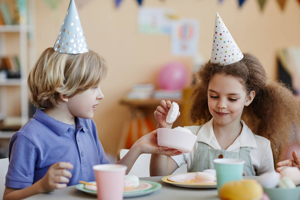 İki mutlu çocuğun portresi parti şapkası takıyor ve masada şekerleme yiyor. - Fotoğraf, Görsel