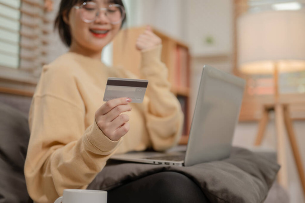 Egy vidám fiatal ázsiai nő regisztrálja a hitelkártyáját egy online vásárlási weboldalon, élvezve az online vásárlást otthon. emberek és életmód fogalmak - Fotó, kép