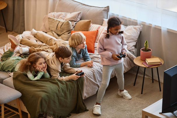 Προβολή υψηλής γωνίας σε ομάδα παιδιών που παίζουν βιντεοπαιχνίδια μαζί και ξαπλώνουν στο κρεβάτι, αντιγραφή χώρου - Φωτογραφία, εικόνα