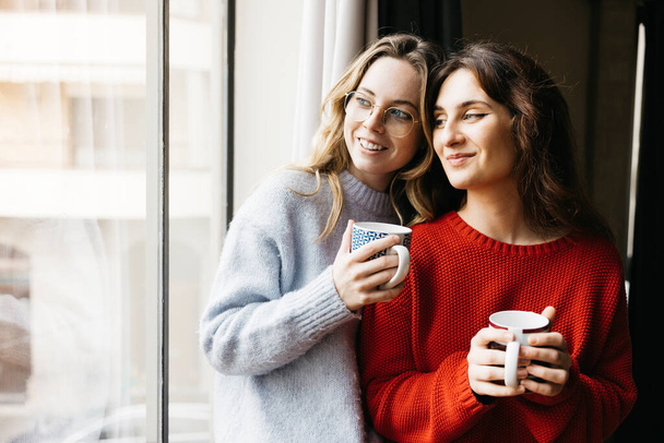 Yanyana duran iki genç kadın, bir bardak çay tutuyorlar, evlerinin penceresinin yanında, samimi ve samimi bir atmosferde bir arkadaşlık anı paylaşıyorlar.. - Fotoğraf, Görsel