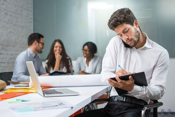 Young businessman holding mobile phone between ear and shoulder talking and writing notes to clipboard in modern office, while his business partners having meeting in the background. - Photo, Image
