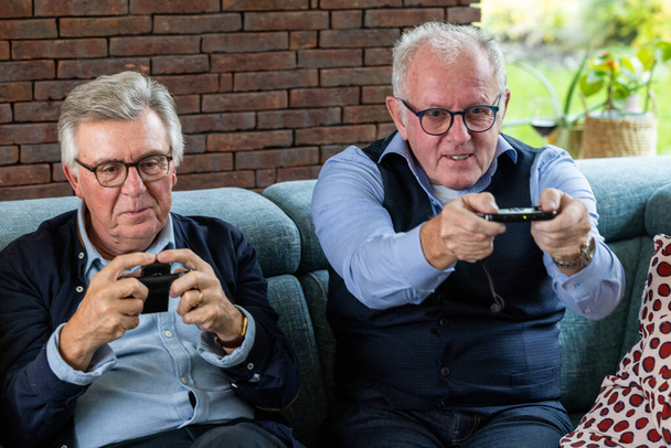 Два старших мужчины проявляют концентрацию и удовольствие, играя в видеоигры, удобно сидя на диване. Кирпичная стена обеспечивает теплый фон, подчеркивая современное времяпрепровождение в домашнем - Фото, изображение