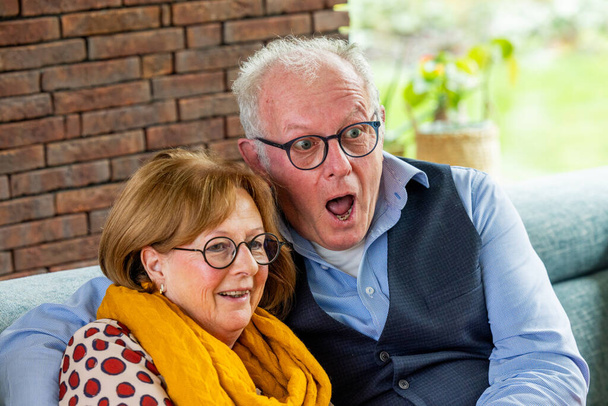Una pareja de ancianos comparte un momento alegre, con el hombre expresando sorpresa mientras la mujer sonríe calurosamente. Se sientan cómodamente en un entorno hogareño, con una pared de ladrillo en el fondo agregando - Foto, Imagen
