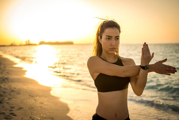 Portrét koncentrované fit mladé sportovkyně oteplování na pláži při západu slunce - Fotografie, Obrázek