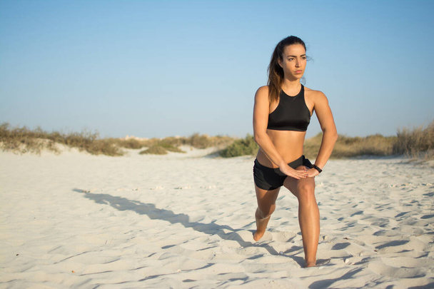 Giovane donna con bel corpo in forma in abbigliamento sportivo stretching gambe con affondo tendine del ginocchio esercizio di stretching sulla spiaggia - Foto, immagini