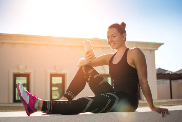 Lächelnde junge Frau in Sportkleidung sitzt auf einer Mauer auf dem Dach eines Gebäudes und benutzt während des Sonnenuntergangs ihr Smartphone - Foto, Bild