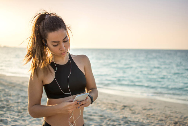 Kulaklıklı genç bir kadının plajdaki akıllı telefondan müzik ve mesaj dinlerken portresi. - Fotoğraf, Görsel