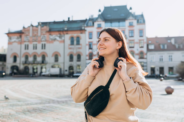 Happy young caucasian woman in good mood with headphones standing on the street in the city. Music lover enjoying music. Urban concept. - Photo, Image