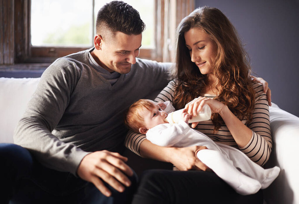 Μητέρα, μωρό και γάλα που τρέφονται με τον μπαμπά σε ένα οικογενειακό σπίτι με αγάπη, υποστήριξη και φροντίδα μαζί. Χαμογελάστε, χαλαρώστε και μπουκάλι για την ανάπτυξη της νεολαίας και την ανάπτυξη με τη μαμά και τον πατέρα σε ένα καναπέ σαλόνι. - Φωτογραφία, εικόνα