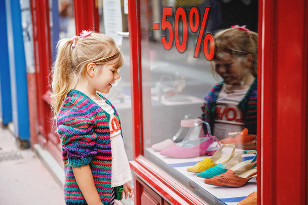 Маленькая девочка смотрит в окно возле торгового центра. Очаровательный счастливый ребенок смотрит на обувь в витрине магазина - Фото, изображение