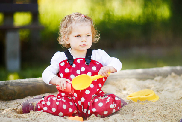 Nettes Kleinkind Mädchen spielt im Sand auf dem Spielplatz im Freien. Schöne Babys in roten Gummihosen haben Spaß an einem sonnigen, warmen Sommertag. Kind mit buntem Sandspielzeug. Gesundes aktives Baby im Freien spielt Spiele - Foto, Bild