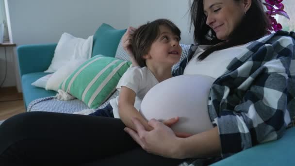 Ragazzo di 5 anni che abbraccia amorevolmente la pancia incinta della mamma sul divano, momento familiare sentito. concetto materno - Filmati, video