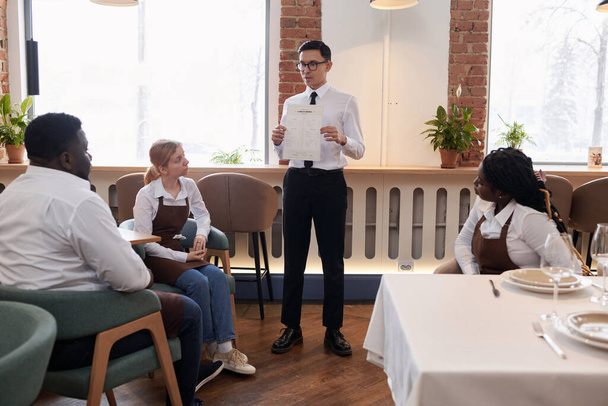 Junger asiatischer Manager arbeitet in modernem Restaurant und zeigt ethnisch unterschiedlichen Bediensteten neue Speisekarte - Foto, Bild
