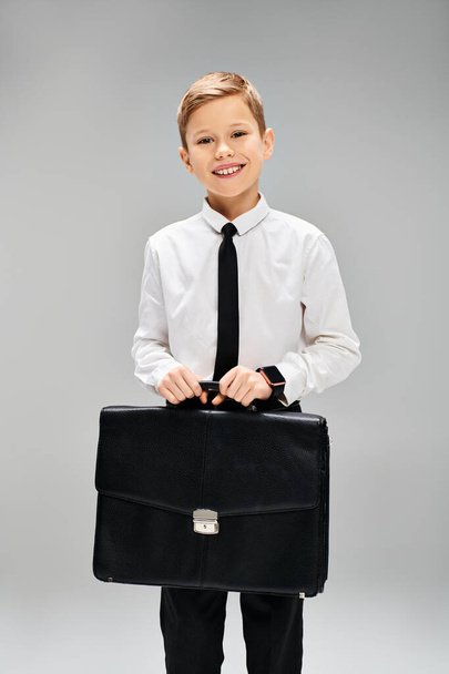Adorable chico de camisa blanca y corbata sosteniendo un maletín negro sobre un fondo gris. - Foto, imagen