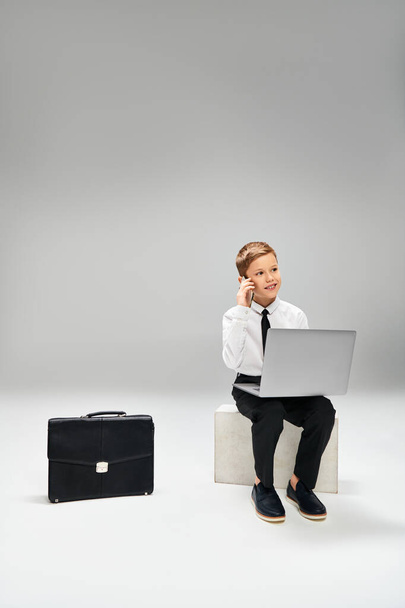 エレガントな服装の若い男の子は,ラップトップに囲まれたスツールに座っています. - 写真・画像
