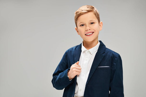 Pieni teini-ikäinen poika sinisessä takissa ja valkoisessa paidassa, huokuu eleganssia ja hienostuneisuutta harmaalla taustalla.. - Valokuva, kuva