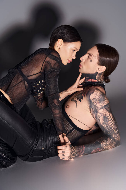 Egy tetovált nő fekszik egy férfi mellett egy stúdióban, szürke háttérrel, egy intimitás pillanatában.. - Fotó, kép