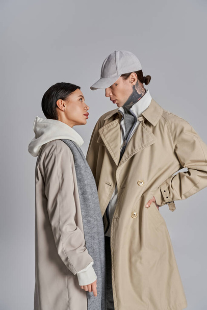 Um jovem casal elegante está lado a lado em casacos de trincheira, exalando sofisticação e charme em um estúdio contra um pano de fundo cinza. - Foto, Imagem