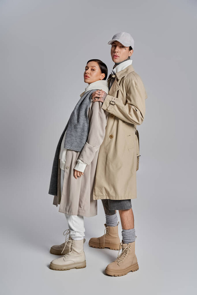 Ein junges stylisches Paar steht Seite an Seite in Trenchcoats, verströmt Raffinesse und Anmut in einem Atelier vor grauem Hintergrund. - Foto, Bild