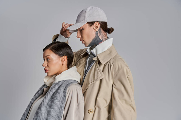 Un joven hombre y una mujer están de pie con estilo en abrigos de trinchera, exudando elegancia y unidad sobre un fondo gris estudio. - Foto, Imagen