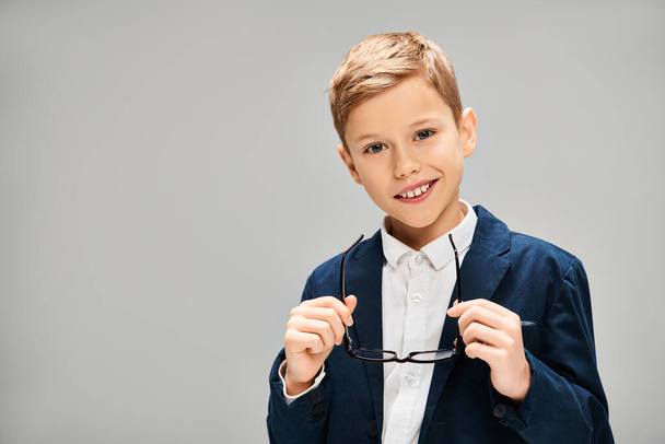 Молодой мальчик в элегантной одежде держит пару очков на сером фоне. - Фото, изображение