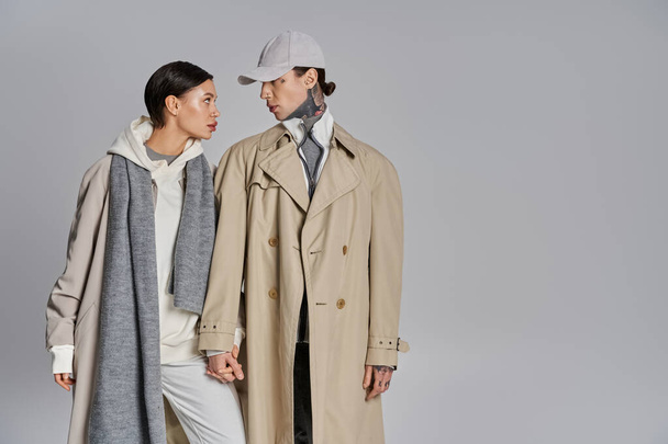 Um jovem e uma mulher estão lado a lado em casacos de trincheira, exalando elegância e estilo em um cenário de estúdio cinza. - Foto, Imagem