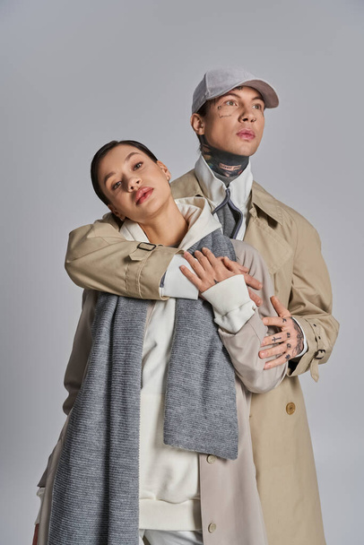 Ein junger Mann und eine junge Frau stehen anmutig nebeneinander in Trenchcoats, die vor grauer Studiokulisse Raffinesse und Stil verströmen. - Foto, Bild