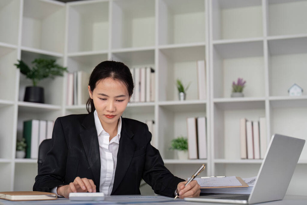 若いビジネス女性アジアまたは従業員会計簿記文書は,ノートパソコンでオフィスで働く財務データやマーケティングレポートをチェックします. ペーパーワーク管理. - 写真・画像