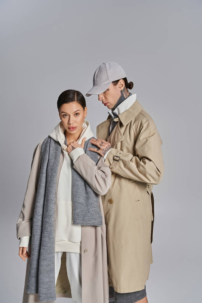 Un giovane uomo e una donna in piedi fianco a fianco, trasudante stile in trench su uno sfondo grigio studio. - Foto, immagini