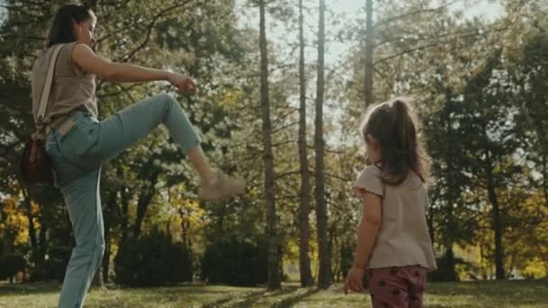 Mutter amüsiert sich mit ihrer Tochter, zeigt ihr, wie man einen Tannenzapfen kickt, spielt gemeinsam im Sommerpark. Konzept einer glücklichen Kindheit. Eine glückliche Familie ist - Filmmaterial, Video