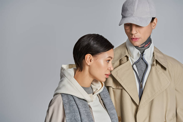 Una pareja joven y elegante en abrigos de trinchera posando con confianza en un estudio sobre un fondo gris. - Foto, Imagen