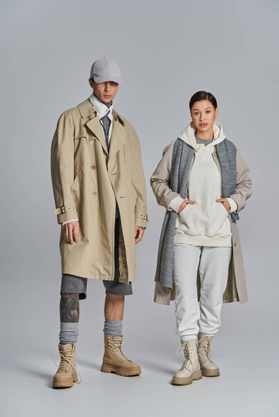 Ein junges stylisches Paar in Trenchcoats steht nebeneinander in einem Atelier vor grauem Hintergrund. - Foto, Bild