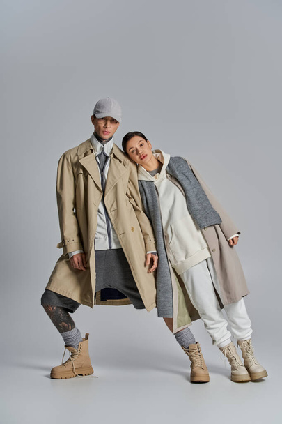 Молода, стильна пара стоїть пліч-о-пліч в траншеї пальто в студії на сірому фоні. - Фото, зображення
