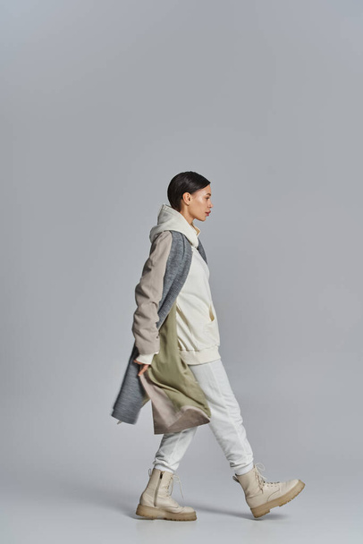 Eine junge, stylische Frau läuft selbstbewusst im Trenchcoat in einem Studio mit grauem Hintergrund. - Foto, Bild
