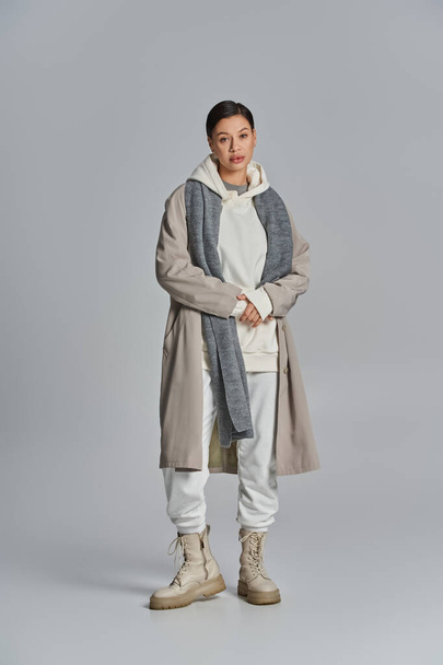 Een jonge stijlvolle vrouw staat vol vertrouwen in een trench jas tegen een strakke grijze achtergrond. - Foto, afbeelding