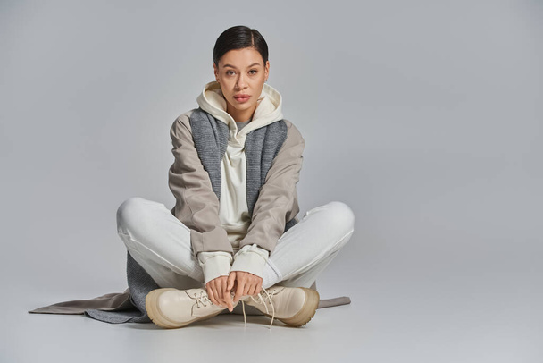 A young stylish woman in a trench coat sits peacefully on the floor, legs crossed, lost in thought in a studio setting with a grey background. - Photo, Image