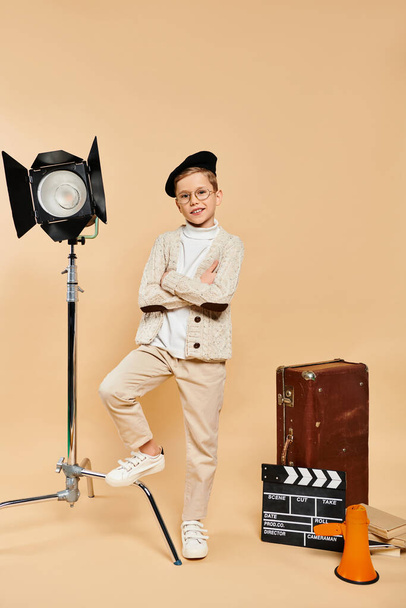 Ένα χαριτωμένο προεφηβικό αγόρι ντυμένο σαν σκηνοθέτης, στέκεται με αυτοπεποίθηση μπροστά σε μια κάμερα.. - Φωτογραφία, εικόνα