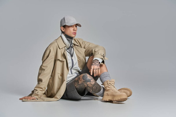 Un joven hombre tatuado en una gabardina se sienta en el suelo con las piernas cruzadas, profundamente pensadas, contra un fondo gris del estudio. - Foto, Imagen