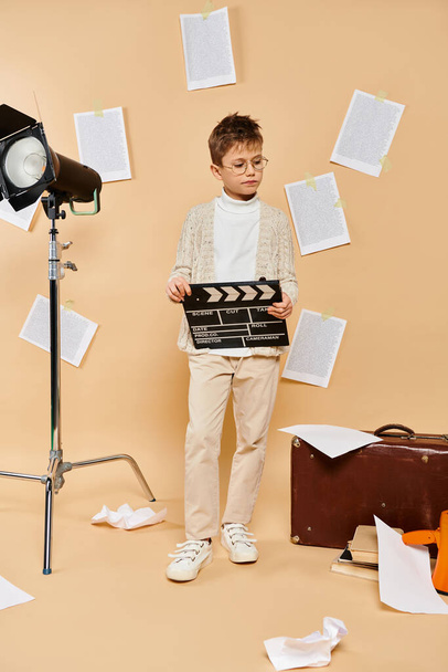 Preadolescent boy holds movie clapper in front of camera on beige backdrop. - Photo, Image