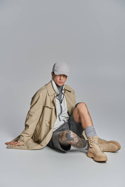 Een jonge, getatoeëerde man in een regenjas zittend op de vloer met zijn benen gekruist in een studio op een grijze achtergrond. - Foto, afbeelding