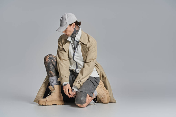 Un jeune homme tatoué assis par terre, coiffé d'un chapeau et d'un trench coat, sur fond de studio gris. - Photo, image