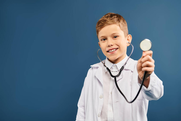 Preadolescent boy in doctors coat holds stethoscope against blue backdrop. - Photo, Image