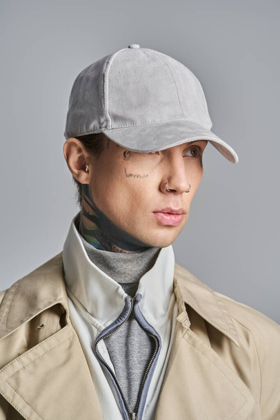 Ein junger, tätowierter Mann strahlt Coolness in einem stylischen Trenchcoat samt schickem Hut vor grauer Studiokulisse aus.. - Foto, Bild