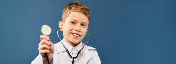 Préadolescent garçon ludique prétendant être un médecin, tenant un stéthoscope sur un fond bleu. - Photo, image
