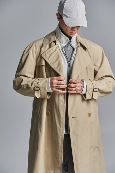 Um homem jovem, tatuado exala mistério e estilo em um casaco de trincheira e chapéu contra um pano de fundo cinza. - Foto, Imagem