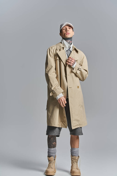 Ein junger, tätowierter Mann posiert selbstbewusst im Trenchcoat in einem Studio vor grauem Hintergrund. - Foto, Bild